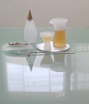 Handcrafted glass teaware by Multeo, including a frosted glass carafe, cup, and tea caddy, arranged with loose tea leaves and a metal tea tool on a reflective glass table.